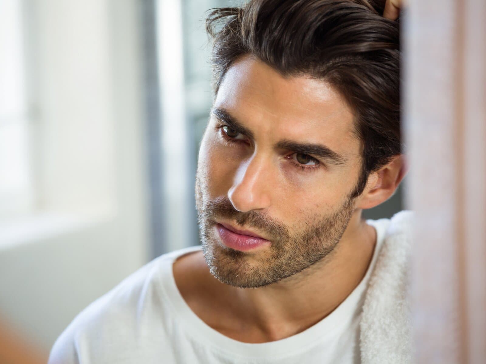 A man running one hand back through thick, dark hair, in a white t-shirt by a window