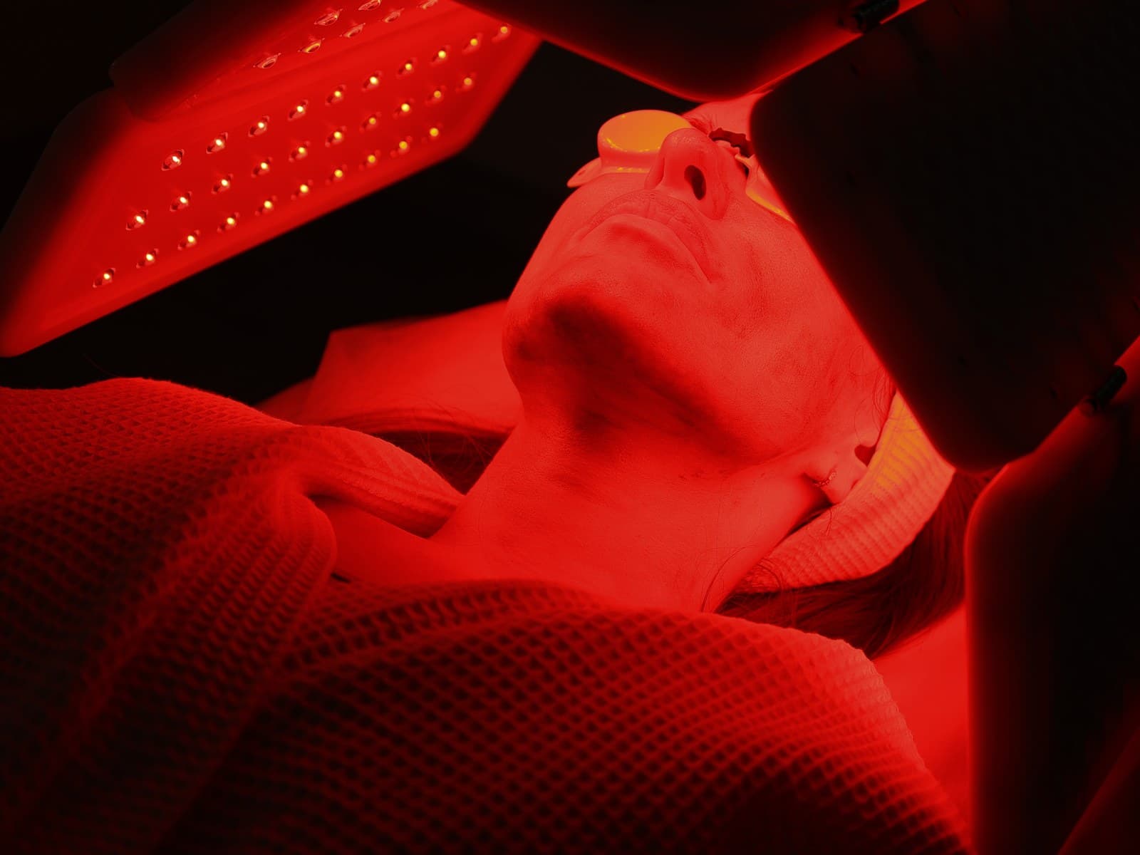 A woman lying back beneath a red LED light panel, her eyes shielded, a waffle blanket drawn to her shoulders