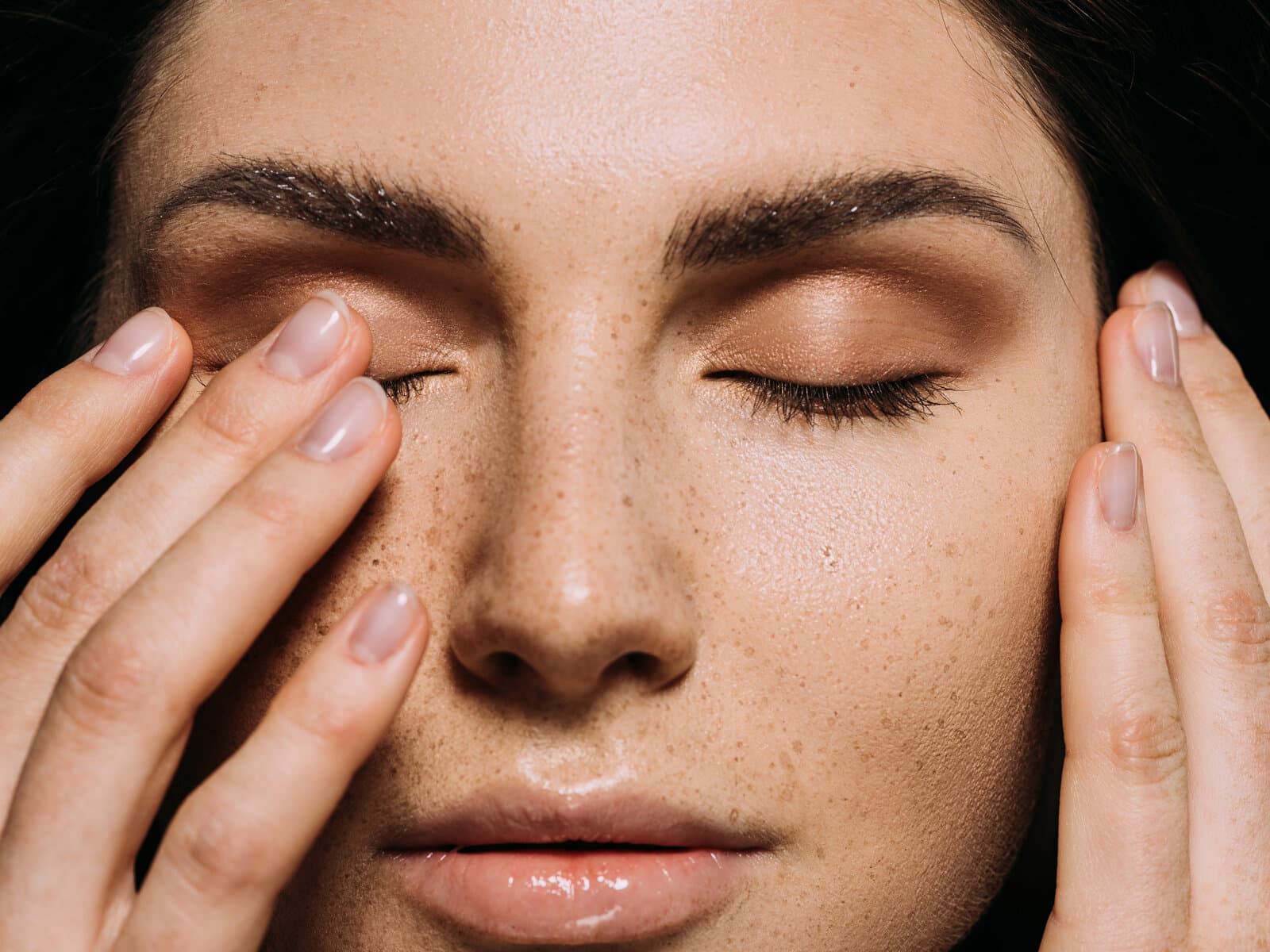 A woman with her eyes closed and her fingertips resting at her temples and cheekbones, freckles across the nose