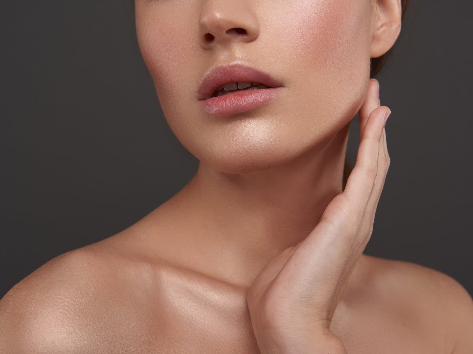 A woman with both hands raised to her jaw, neck and shoulders bare, against a dark ground